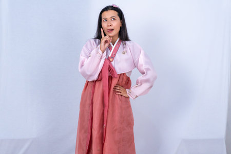 A woman in pink and red Korean Hanbok looks up thinking with a tongue out on a white backgroundの写真素材