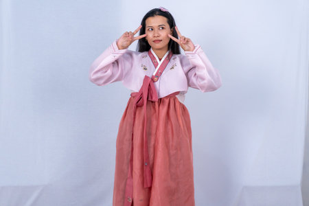A woman in pink and red Korean Hanbok makes peace signs with a smile against a white backgroundの写真素材