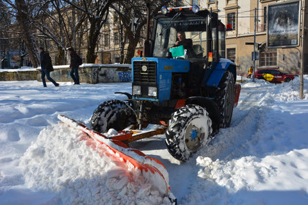 Snow tractor removal with rotating brush sweeping snow from footpath on embankment in park. Red tractor with scoop and automated brush removes snow. Municipal road sweeping vehicle with plowのeditorial素材