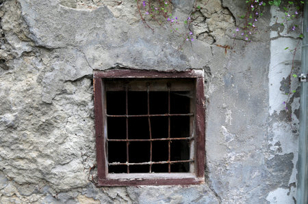 Old window with metal bars set in a stone wall of an abandoned building. Cracked plaster, rough...