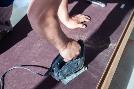 Closeup hand of woodworker with professional cutting tool fretsaw or jigsaw, cut wooden tabletop, sawing plank, brown filings, sawdust. Concept professional service for installation and repairの写真素材