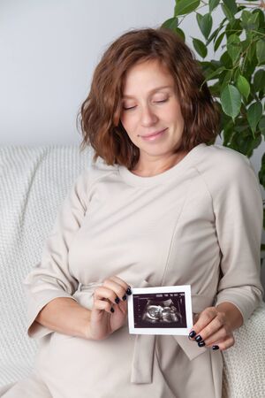 Pregnant woman holding ultrasound image. Concept of pregnancy, health care, gynecology, medicine. Young mother waiting of the baby. Close-up, copy space, indoors.の写真素材