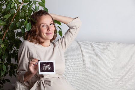 Pregnant woman keeping hand on belly and holding ultrasound image at home interiors. Pregnancy,...
