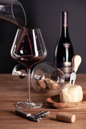 Pouring red wine in crystal glass, bottle, corkscrew, opener, sommelier knife, transparent decanter, corks, cheese on wooden table. Closeup, vertical, side view. Concept degustation winery cellarの写真素材