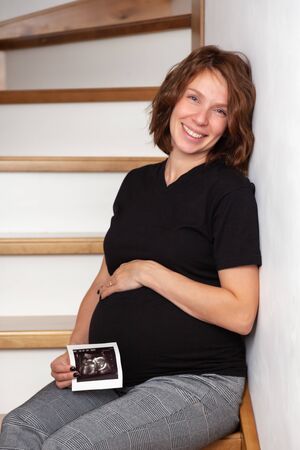 Image beautiful pregnant smiling caucasian curly woman sitting on wooden stairs and posing while showing ultrasound scans on her tummy on white background. Looking at the cameraの写真素材