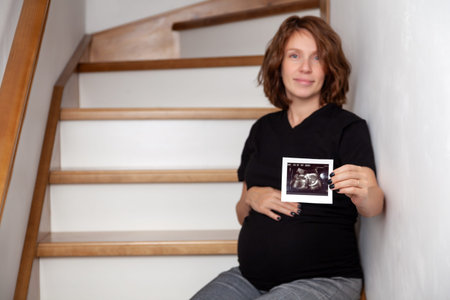 Image of pregnant smiling caucasian curly woman sitting on wooden stairs and posing while showing...
