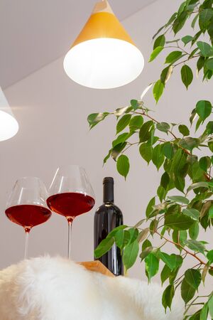 Dark bottle and two glasses of red wine on table in modern kitchen, lamps over table, green leaves of tree. Vertically. Copyspace. Place for text, lettering, advertisement in catalogの写真素材