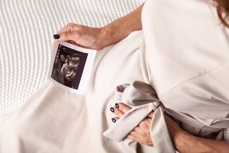 Pregnant woman keeping hand on belly and holding ultrasound image at home interiors. Pregnancy, parenthood, preparation and expectation concept. Close-up, copy space, indoors.の写真素材