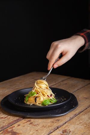Lunch table - closeup male hand with italian spaghetti on vintage fork serving on black dishes. Pasta with vegetables, pepper, basil leaves on wooden background. Verticalの写真素材