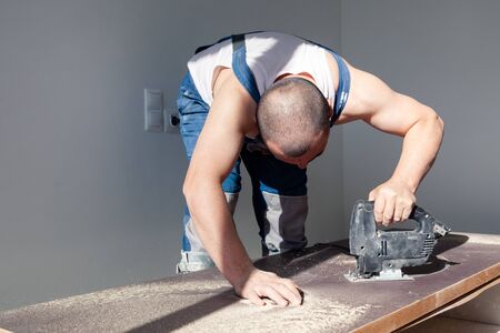Closeup hand of carpenter, woodworker with professional cutting tool fretsaw or jigsaw, cut wooden tabletop, sawing plank, brown filings, sawdust. Concept cut hole for sink in the kitchenの写真素材