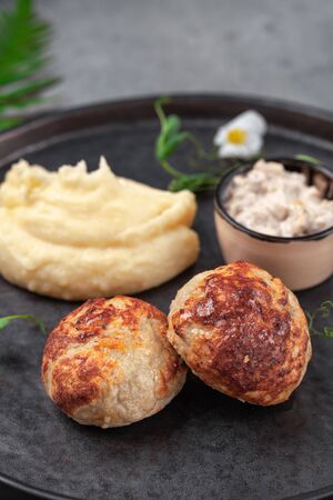 Portion of chicken meat patties with sauce, mashed potatoes, herbs, decorated with flowers on round black plate on gray surface, close-up, view from above, place for text, copy space, verticalの写真素材