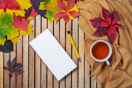 Autumn leaves, white notebook, tea cup on table, textile napkin, wooden background