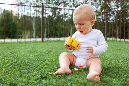 Funny blond cute baby sitting in grass and playing with car toy