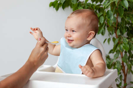 Mother feeds funny happy blond baby in blue bib from a spoonの写真素材