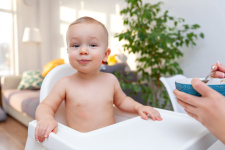 Mothers hand feeds funny cute happy blond baby toddler with spoon