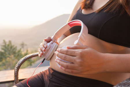 Pregnant woman putting headphones on belly on a chair at mountain sunset time, expectation conceptの写真素材