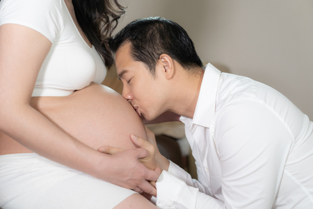 Asian couple pregnant wife and husband kissing on belly woman in bedroom background.の写真素材