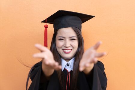 Portrait closeup. Asian beautiful smiley graduate graduated student girl young woman in cap gown on isolated orange background wall. Celebrating graduation ceremony concept.の写真素材