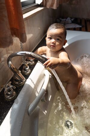 Asian happy laughing baby boy taking a bath playing with foam bubbles. Little child in a bathtub. Smiling kid in bathroom with sunlight window background. Hygiene and care for young children concept.の写真素材