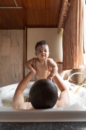 Asian happy baby boy and father playing with foam bubbles. Little child in a bathtub. Smiling kid in bathroom with sunlight window background. Hygiene and care for young children concept.の写真素材