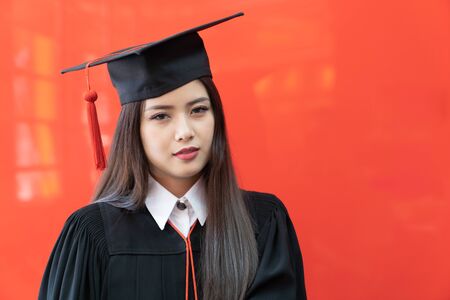 Portrait closeup. Asian beautiful smiley graduate graduated student girl young woman in cap gown on isolated red background wall. Celebrating graduation ceremony concept.の写真素材