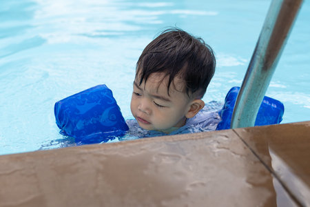 Asian cute little toddler boy in a colorful swimming suit relaxing in a pool having fun during...