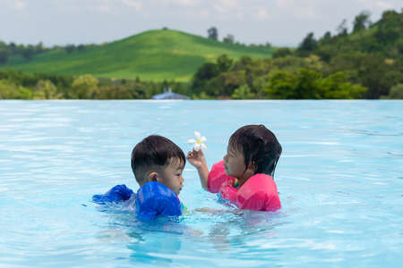 Asian cute little toddler boy and girl in swimming suit relaxing in a pool having fun during summer vacation in a tropical resort, mountain background.の写真素材
