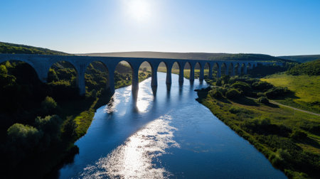 A majestic viaduct arches gracefully over a calm river, surrounded by lush green hills under a clear blue sky. The sunlight glints off the water's surface, enhancing the tranquil beauty.の素材