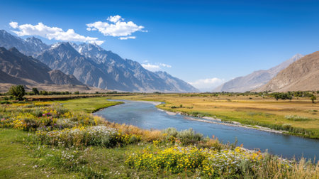 A tranquil river winds through a vibrant valley filled with wildflowers, while towering mountains rise in the background under a bright blue sky.の素材