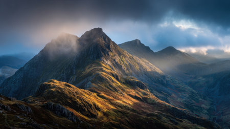 Sunlight breaks through clouds illuminating the rugged mountain peaks and lush valleys below.の素材