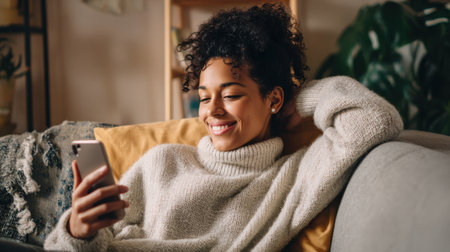 A woman with curly hair enjoys her time on a couch, smiling as she interacts with her smartphone. She is dressed in a warm sweater, surrounded by plants and soft lighting.の素材