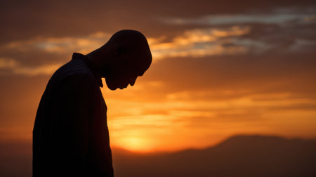 A person stands with a bowed head against a vibrant sunset background. The warm colors of the sky create a peaceful atmosphere, encouraging moments of reflection and tranquility.の素材