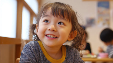 A young girl with curly hair smiles widely while sitting at a table in a warm, inviting classroom. Other children can be seen engaged in various activities around her.の素材