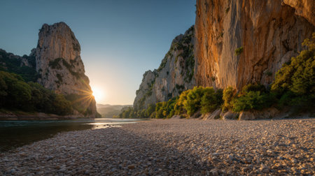 The sun sets behind towering rock formations, casting a warm golden glow over the river. Lush trees line the banks, adding life to the tranquil canyon scene.の素材
