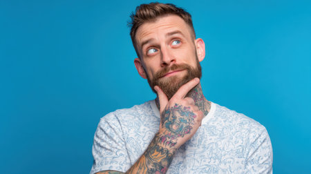 A man with a beard and tattoos rests his chin on his hand while looking up thoughtfully. The bright blue backdrop enhances his casual attire and expressive pose.の素材