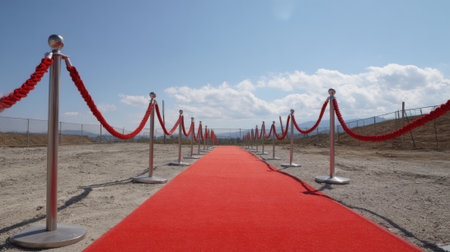 A vibrant red carpet stretches across a dry construction site lined with elegant stanchions and ropes. Clear skies and distant hills create a promising backdrop for an upcoming event.の素材