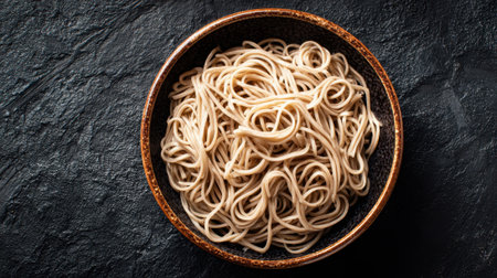 A bowl filled with freshly prepared soba noodles is placed on a dark, textured surface. The noodles are long, thin, and arranged in a tangled manner, inviting a culinary experience.の素材