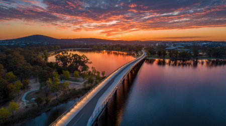 A stunning sunset casts vibrant colors across the water as a winding path appears alongside. Trees line the shore creating a peaceful scene near the city.の素材