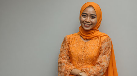 A woman wearing a vibrant orange dress and hijab smiles warmly posing gracefully with her arms crossed.の素材