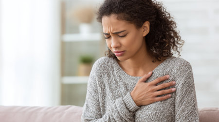 A woman sits on a sofa clutching her chest with a pained expression amidst a warm living room.の素材