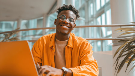 A cheerful young man sits at a table using a laptop. He wears an orange jacket and glasses. Bright sunlight fills the spacious modern workspace.の素材