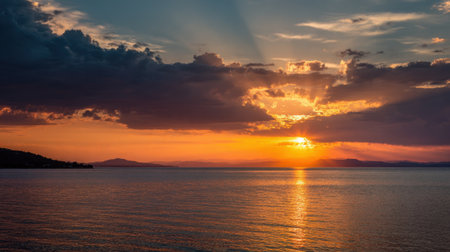 Vibrant sunset paints the sky in shades of orange and purple over a serene lake. Silhouetted hills create a peaceful backdrop as gentle waves ripple in the water.の素材