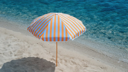 A vibrant orange and white striped beach umbrella is placed on the sandy beach. Gentle waves lap at the shore under a clear blue sky during a sunny day, creating a perfect seaside scene.の素材