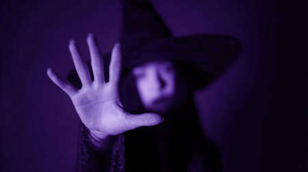 A witch dressed in dark attire extends her hand forward in a magical gesture. The purple background adds an eerie atmosphere enhancing the mystic aura of the scene.の素材