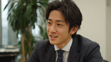 A young man with dark hair smiles while speaking in a professional office setting during the daytime.の素材