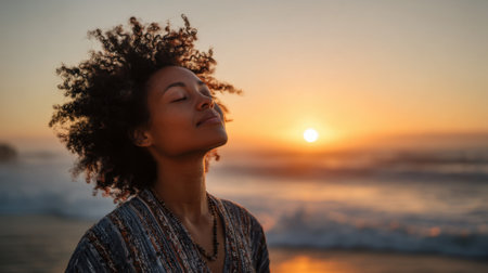 A woman stands on the beach with her eyes closed savoring the calmness of the ocean waves as the sun sets on the horizon. Her hair is illuminated by warm sunset colors.の素材