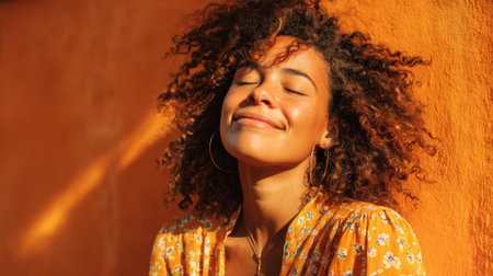 A young woman relaxes with her eyes closed basking in the sunshine. She has curly hair and is wearing a floral dress. The warm orange wall adds a cheerful backdrop to the peaceful scene.の素材