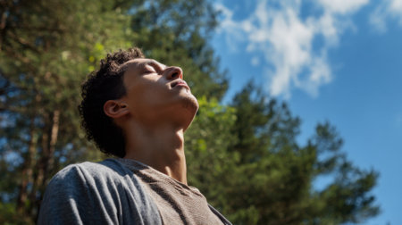 A young man stands outside with his eyes closed taking in the fresh air. He appears relaxed and at peace while surrounded by tall trees and bright blue skies.の素材