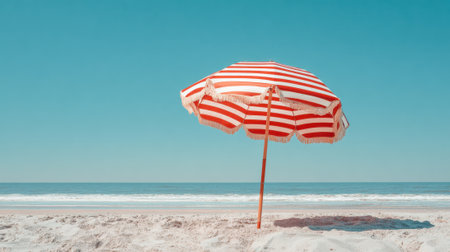 A colorful umbrella stands alone on the sandy shore, casting a shadow on the beach. The ocean waves gently lap against the shoreline, and the sky is clear and blue.の素材