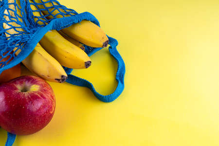 Blue bag string bag with bananas, red apples and tangerines on a bright yellow background. Concept of eco-friendly materials. Close up. Copy space.の写真素材